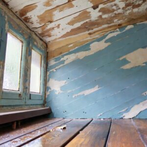 A close-up of a water-stained ceiling, peeling paint, and warped wooden floor